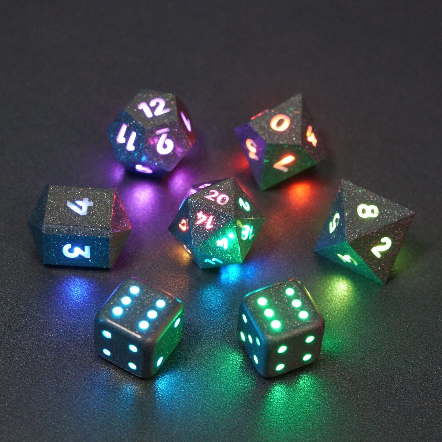 Set of 7 lit Hematite Grey dice with a rainbow of colors across all faces. Set Includes: 1 D20, 1 D12, 1 D10, 1 D8, 2 Pipped D6, 1 D4. Hematite Grey colorway is a fully opaque medium tone silver resin packed with rainbow glitter of various sizes. The numbers or symbols are painted pale white.