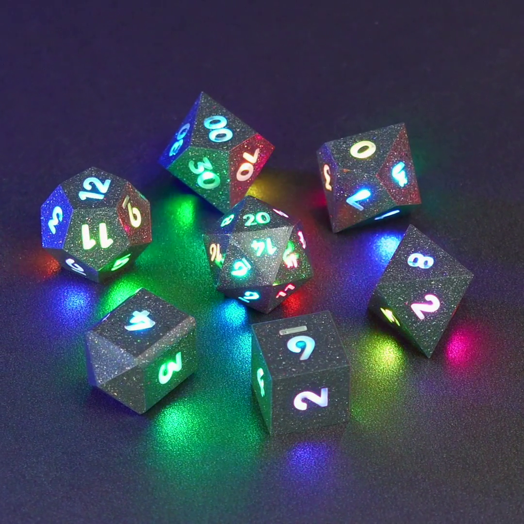 Set of 7 lit Hematite Grey dice with a rainbow of colors across all faces. Set Includes: 1 D20, 1 D12, 1 D00, 1 D10, 1 D8, 1 D6, 1 D4. Hematite Grey colorway is a fully opaque medium tone silver resin packed with rainbow glitter of various sizes. The numbers or symbols are painted pale white.