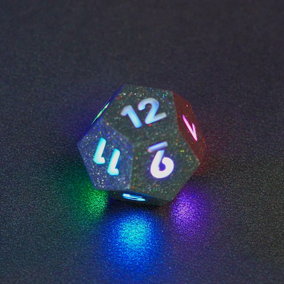 Lit Hematite Grey D12 with a rainbow of colors across each face. Hematite Grey colorway is a fully opaque medium tone silver resin packed with rainbow glitter of various sizes. The numbers or symbols are painted pale white.