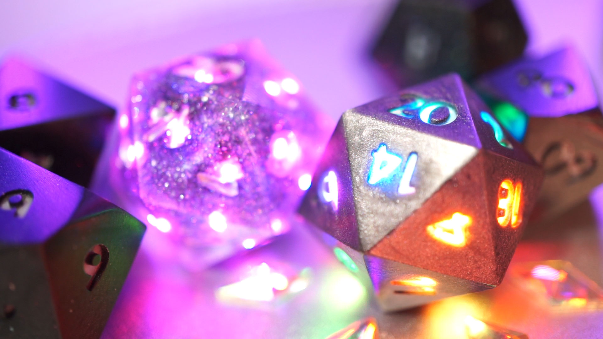 Photograph of multiple dice with two d20s in the center as the focus. One is filled with glitter and has purple colored LEDs lighting up all translucent faces, the other is opaque metallic gray and has different color LEDs lighting up individual numbers on each face.