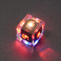 Lit Clear D6 with red and blue colors across each face. Clear colorway is fully transparent resin allowing internal circuit board to be visible. The numbers or symbols are painted metallic copper.