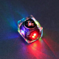 Lit Clear D4 in a crystal shape with a rainbow of colors across each face. Clear colorway is fully transparent resin allowing internal circuit board to be visible. The numbers or symbols are painted metallic copper.