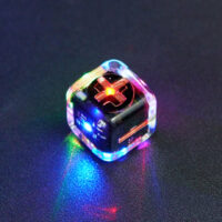 Lit Clear Fudge D6 with a rainbow of colors across each face. Clear colorway is fully transparent resin allowing internal circuit board to be visible. The numbers or symbols are painted metallic copper.