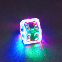Lit Clear Pipped D6 with a rainbow of colors across each face. Clear colorway is fully transparent resin allowing internal circuit board to be visible. The numbers or symbols are painted metallic copper.